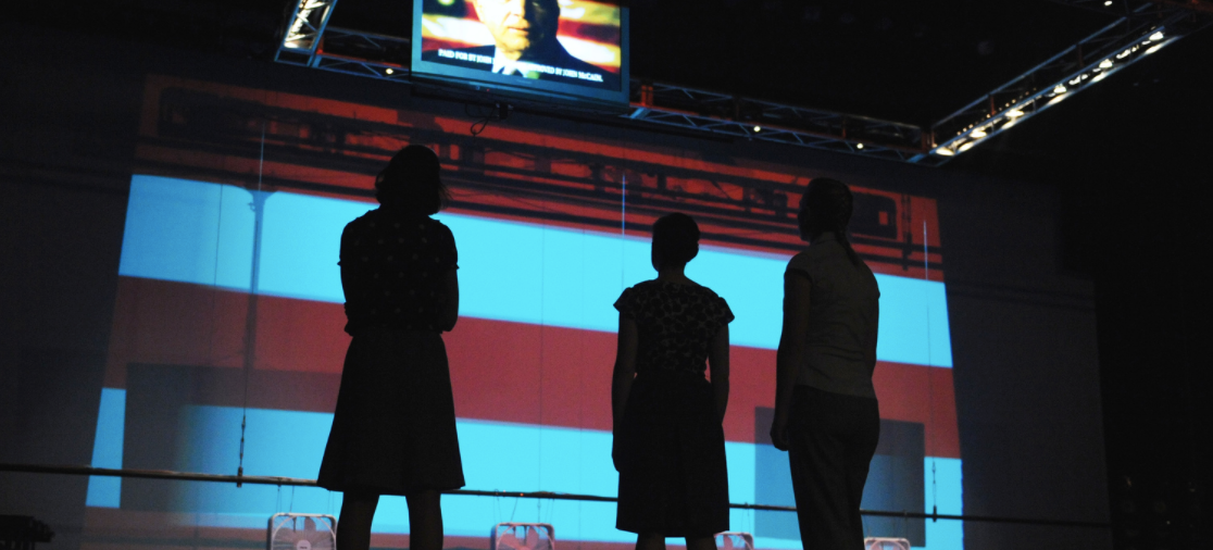 Silhouette of three students against the backdrop of the American flag, looking up at a screen of election coverage in the 2008 production of The Race at Georgetown University.